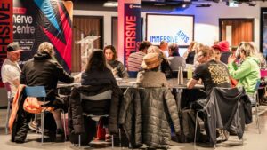 A diverse group of people sits around a table engaged in discussion at an event. Banners and signs reading "Immersive" and "Nerve Centre" are visible.
