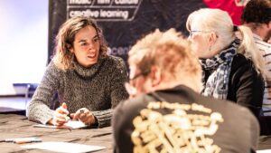 A woman smiling and talking with others at a table, showing engagement. Papers and pens lie before her. The setting feels collaborative and positive.