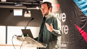 Tom a man with glasses speaks at a podium with a laptop, gesturing with one hand. The background features a dark banner with partial text visible, creating a professional and engaged atmosphere.