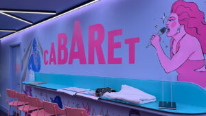 Vibrant cabaret-themed lounge with pink and blue decor. A mural features a performer singing passionately. Pink chairs line a counter with magazines.