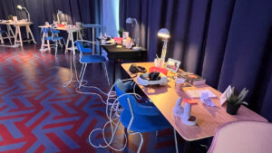 A room with dim lighting features several wooden tables with blue chairs. Each table has table lamps, books, and virtual reality headsets, creating a tech-focused, cozy atmosphere.