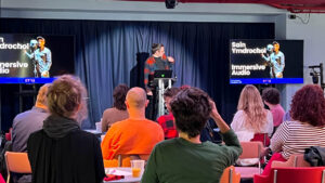 A presenter stands at a podium in front of an audience, gesturing towards slides on dual screens labeled "Sain Ymdrochol" and "Immersive Audio."