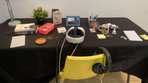 A desk with a black tablecloth holds books, framed photos, a plant, and various office supplies. A white headphone set is centred. A yellow chair is in front.