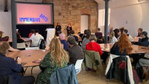 A group of people sits attentively at round tables in a room with exposed brick walls. A large screen displays "Immersive Arts" as two speakers present.