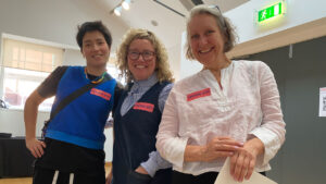 Three female Immersive Arts producers are smiling, each wearing an "Immersive Arts" sticker. They're standing indoors, conveying a cheerful and welcoming atmosphere.