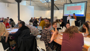 A lively workshop setting with people seated at tables, engaged in discussion. A presentation is visible on a screen, creating an educational atmosphere.