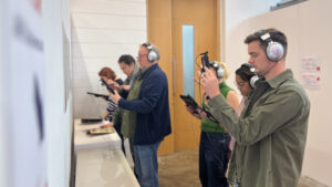 Five people in a line, wearing headphones, engage with handheld devices in an indoor setting. The mood is focused and attentive.