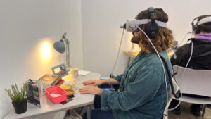 A person with a beard wearing a denim shirt sits at a desk using a VR headset. The desk has a lamp, plant, books, and open boxes, creating a focused atmosphere.