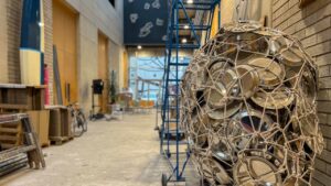 Art installation in a modern indoor space with a sculpture of metal lids tangled in rope. A blue ladder and scattered furniture enhance the creative atmosphere.