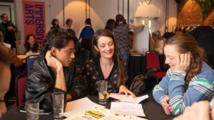A group of three people sit at a table in a lively workshop or event space. They are engaged in discussion, surrounded by papers and drinks. A poster reads "Immersive Arts" in the background. The atmosphere is collaborative and creative.