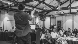 Tom Millen presents to an attentive audience in a modern industrial-style room. The black-and-white image conveys an educational and engaging atmosphere.