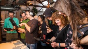 A group of seven people in casual attire are using tablets and smartphones in a lively indoor setting. They appear engaged and focused, smiling and sharing information.