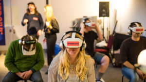 A group of people wearing virtual reality headsets labeled "Immersive Arts" sit in a room, engaged and focused. The atmosphere is interactive and exploratory.