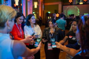 A group of people at a lively event, smiling and holding smartphones, wearing lanyards. The room is warmly lit with colorful lights and a large screen.