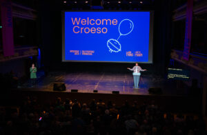 A person speaks at a podium on a stage with a large screen displaying "Welcome Croeso" and graphics, addressing a seated audience in a dimly lit theater.