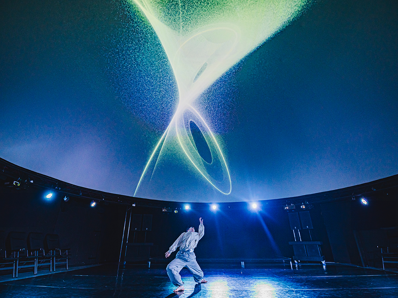 A dancer stands alone on a dark, circular stage, mid-movement with one arm reaching upward. Above them, a vast curved screen or dome fills the space with swirling, luminous abstract light patterns in blues, greens and soft whites, resembling cosmic energy or flowing particles. Stage lights line the edge of the space, and the scale of the projection dwarfs the performer, creating a sense of awe, immersion and connection between the human body and an expansive digital environment.