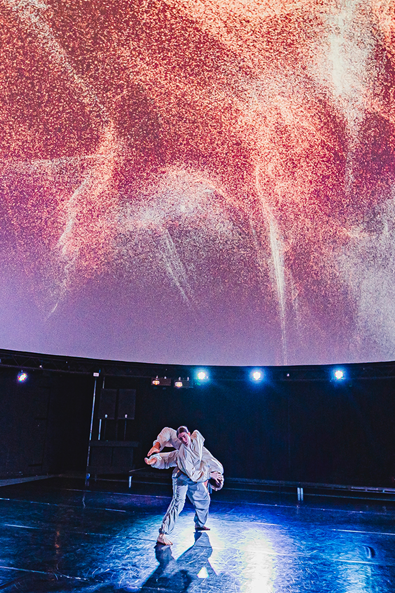 A lone dancer stands on a dark, glossy stage beneath a large domed ceiling filled with abstract, cosmic-like projections in red, purple, and white tones, resembling swirling stars or dust.