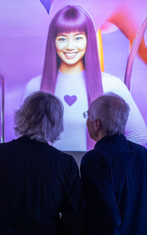A couple is facing away from the camera in shadow. They are looking ahead at a large projection of an AI generated image of a smiling woman with long pinky/ purple hair and a lilac top with a heart on it.