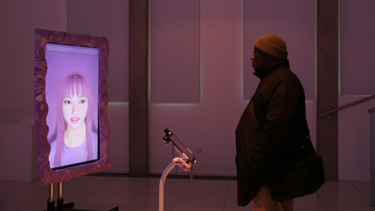 A man looks at a large monitor on a stand facing him. The screen shows a pink background and an AI women with pink hair. The screen has a squidgy pink frame.