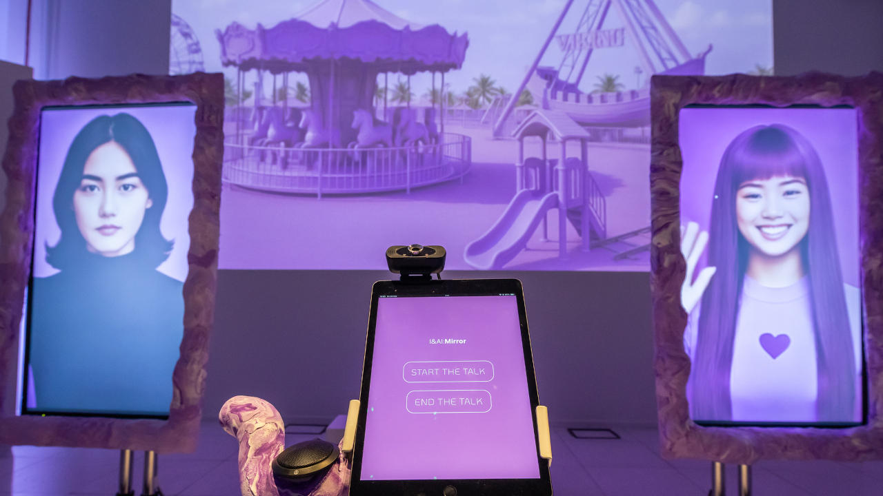 A tablet is at the base of the screen with two buttons, one to start and one to end the talk. There are two large monitors in the background, one on each side and right at the back is a large projection. The two monitors have a face on each, the left monitor has an AI woman with short brown hair and a black top, whilst the right monitor an AI woman with long purple hair waving and a lilac top with a heart on it. The projection at the back shows a dreamy pink funfair AI landscape.