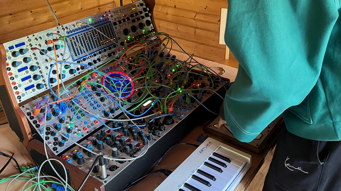 Sonia is wearing a big turquoise jumper and standing over a modular synthesiser with lots of colourful wire outputs, and a keyboard sits below it.