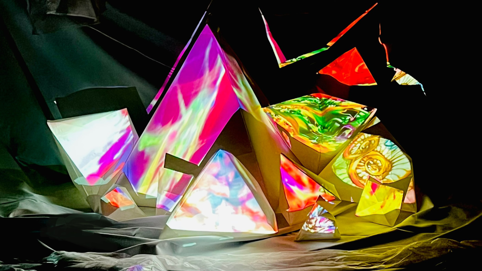An abstract installation made from folded, angular sculptural forms arranged like crystalline peaks. The surfaces are illuminated with vivid projected colours—pinks, greens, yellows, blues, and swirling organic patterns—creating the effect of glowing prisms in a dark space. The structures sit on soft, draped fabric, with sharp edges and layered planes catching the light,