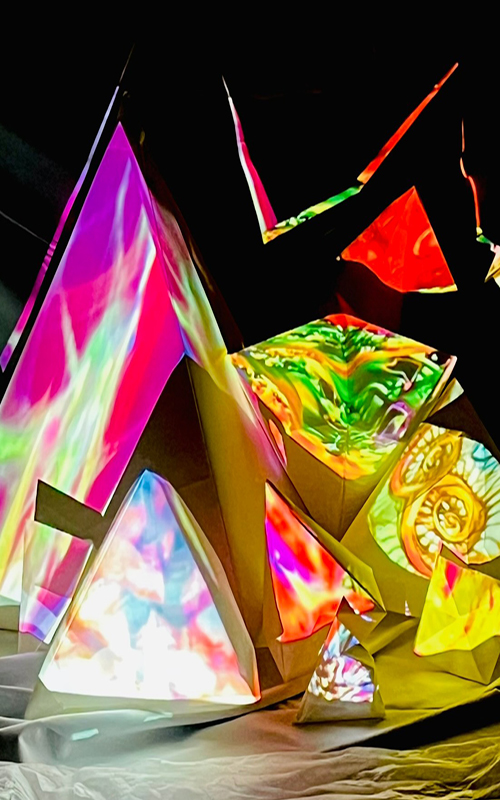 An abstract installation made from folded, angular sculptural forms arranged like crystalline peaks. The surfaces are illuminated with vivid projected colours—pinks, greens, yellows, blues, and swirling organic patterns—creating the effect of glowing prisms in a dark space. The structures sit on soft, draped fabric, with sharp edges and layered planes catching the light,