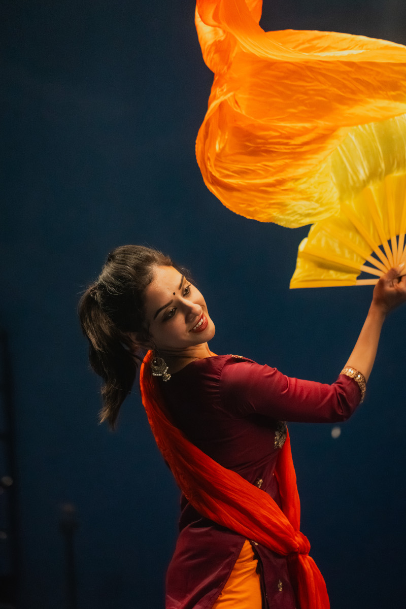 South Asian dancer wearing a red long sleeved dress. The dancer is waving a yellow fan and there is a long billowing orange scarf.