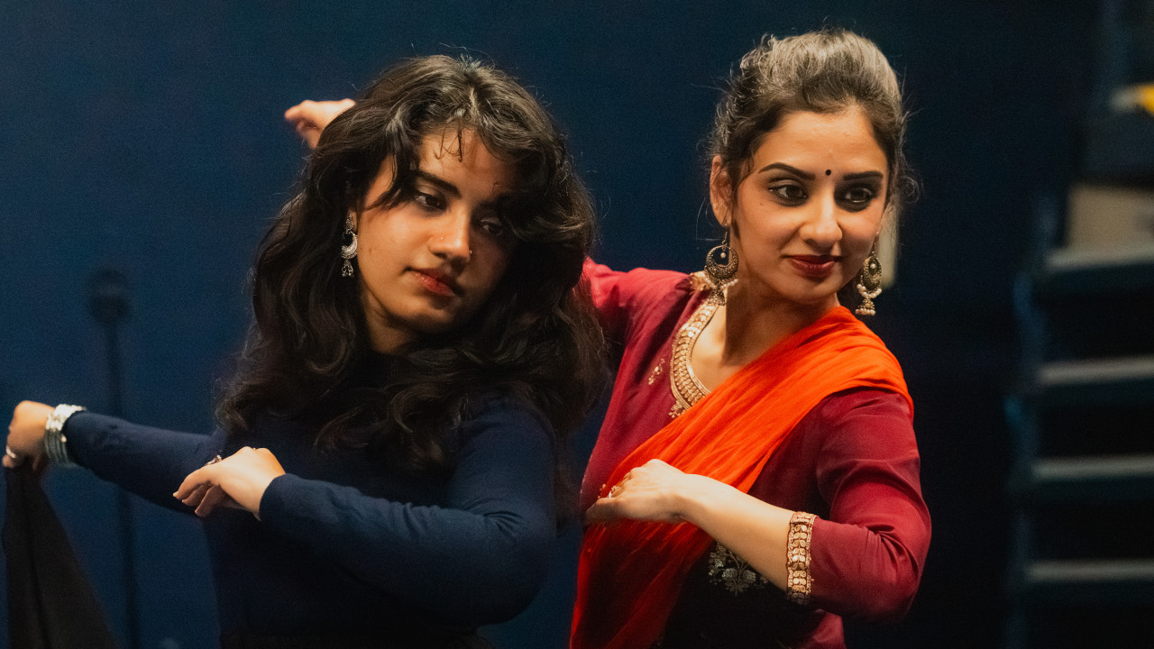 Two south asian women practicing a dance. They are facing forwards and looking slightly down with their arms facing behind them.