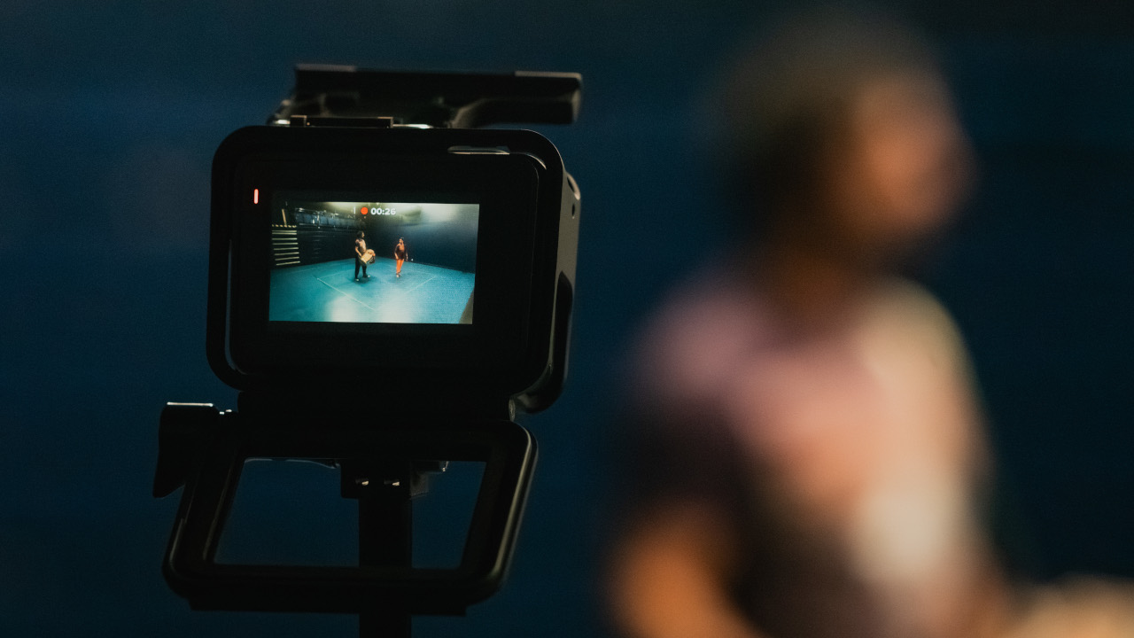 a video camera is in the foreground but the background is blurry. The videocamera is capturing someone dancing in an large empty rehearsal space.
