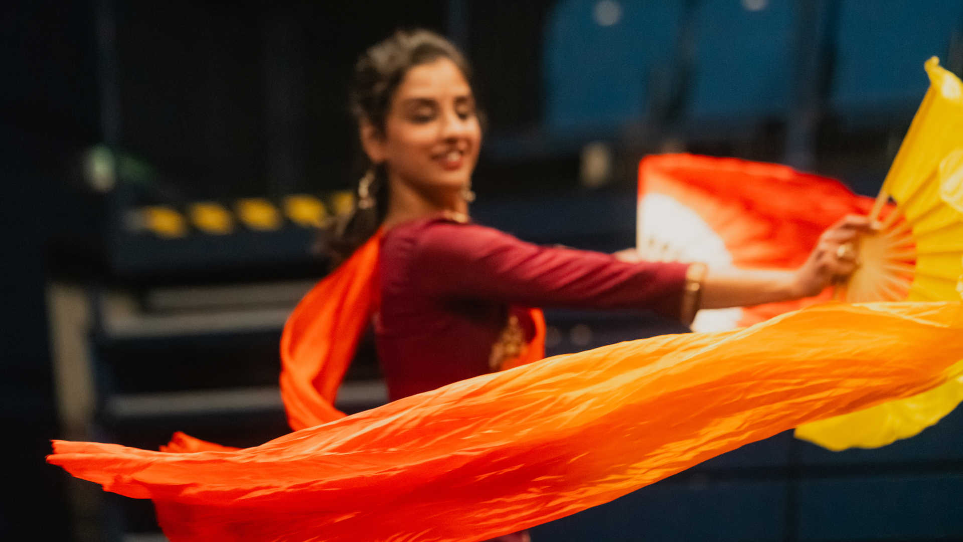 South Asian dancer wearing a read long sleeved shirt and wave an orange and yellow fan in each hand, alongside a long billowing scarf in fiery orange and reds