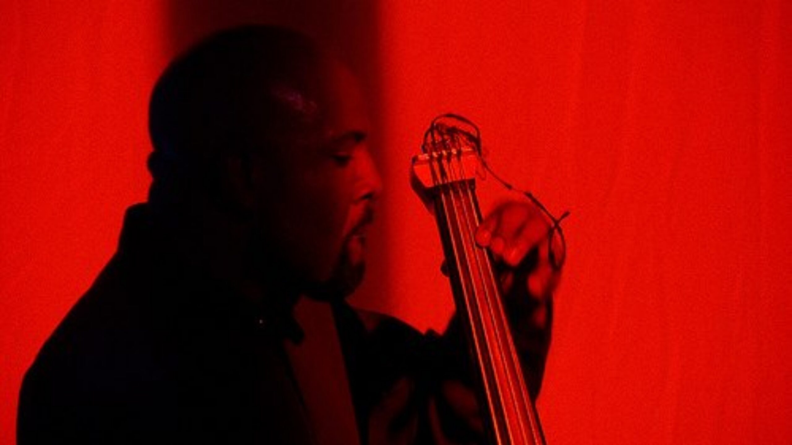 A Black person plays an upright bass against a vibrant red background, creating a dramatic and intense atmosphere. The focus is on the musician's concentration.