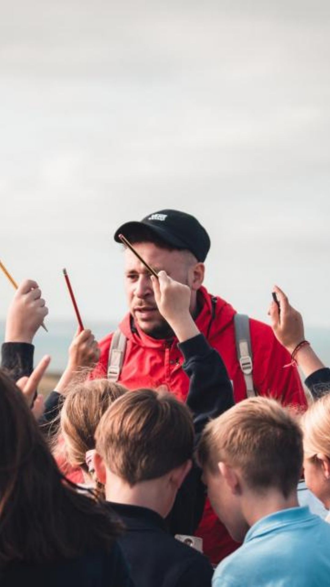 A man in a red jacket and black cap surrounded by children holding colourful pencils aloft, creating a lively and inspiring outdoor scene.