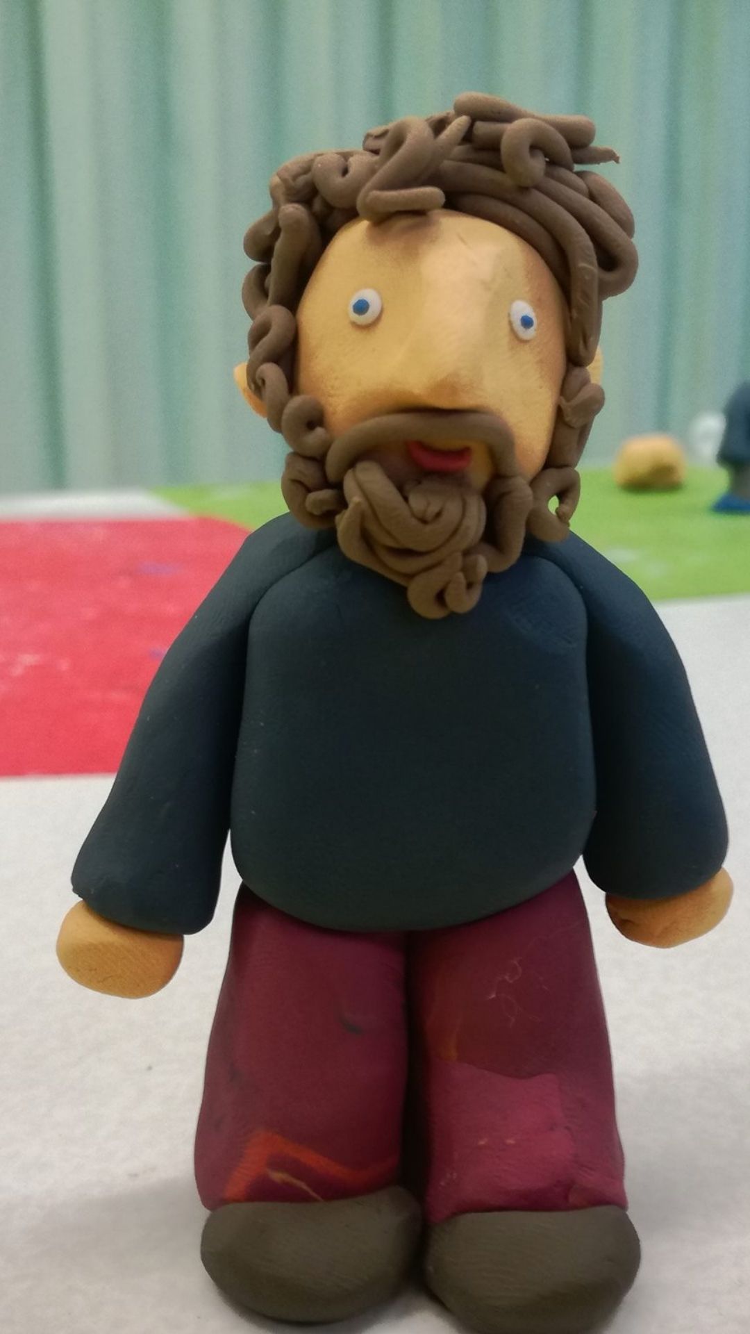 Clay figure of a person with curly hair and a beard, wearing a dark shirt and red pants. It's placed on a table with a colorful, blurred background.