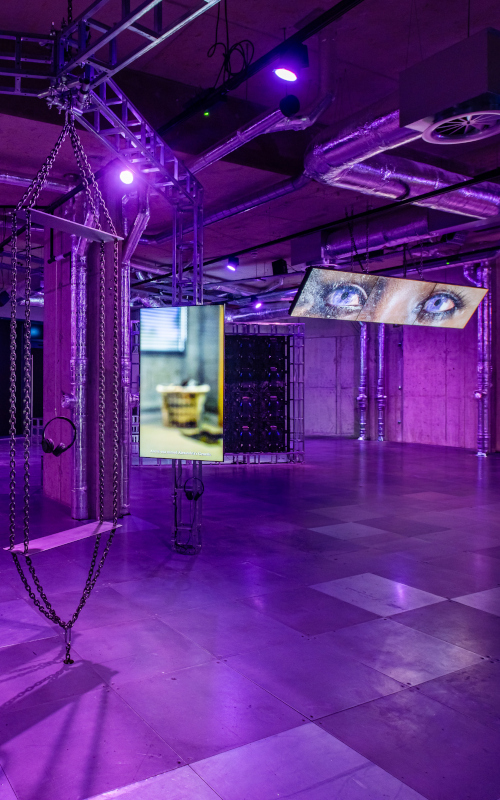 exhibition installation with monitors across a large industrial room lit in purple light