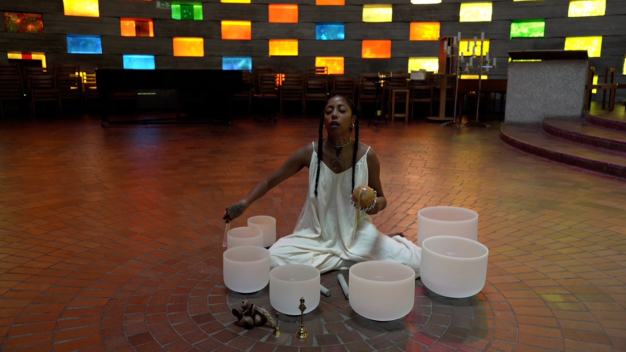 Frances Young_EXPLORE artist_photo of a woman sitting on the floor surrounded by singing bowls against a background of glass bricks in multiple colours