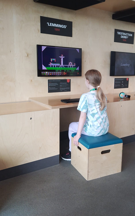 a child engages with retro video games at a museum, one seated, focused on "Lemmings,"