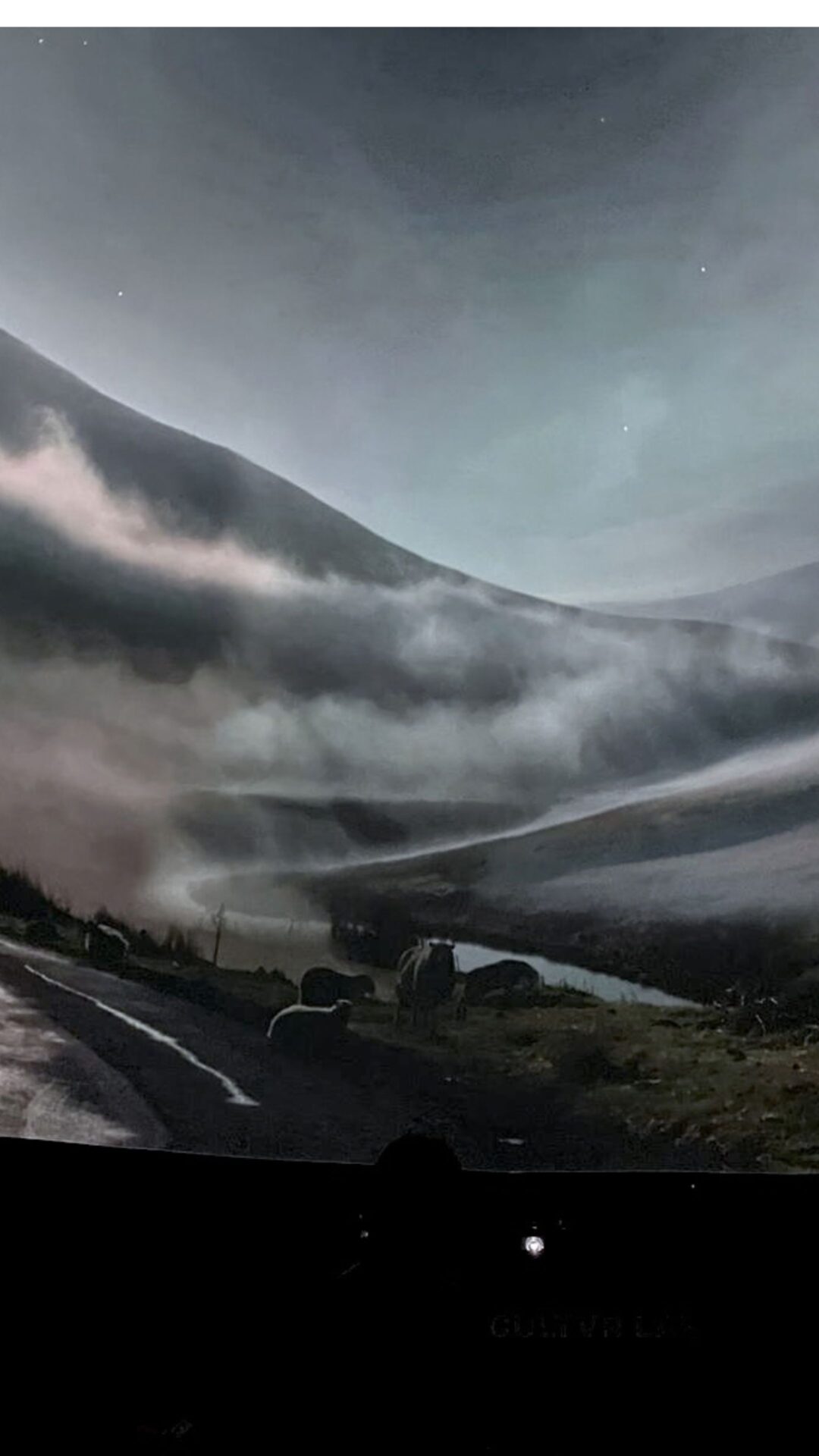 A dramatic, mist-filled mountain valley surrounds a winding road beneath a vast gray sky, creating a moody, cinematic sense of isolation and awe.
