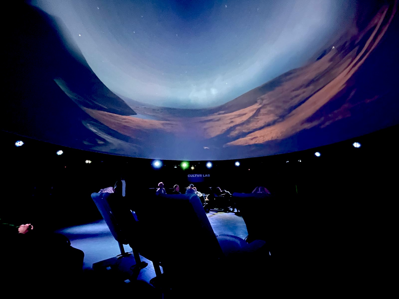 A dark planetarium theater with a sweeping, curved screen showing a blue sky above a rugged desert canyon; seated viewers watch in a quiet, immersive atmosphere.
