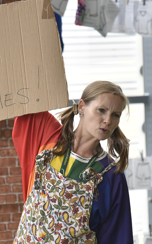 Someone with blonde hair, a rainbow parachute top, and floral dungarees stands defiantly with a sign saying 'NO BALL GAMES' - the 'NO' is crossed out.