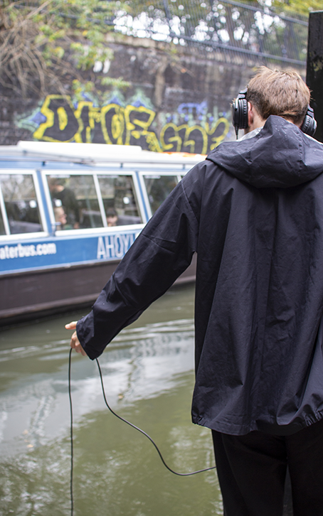 A person wearing headphones stands by a graffiti-covered wall, facing a blue canal boat. The scene has an urban, casual vibe with a rainy atmosphere.