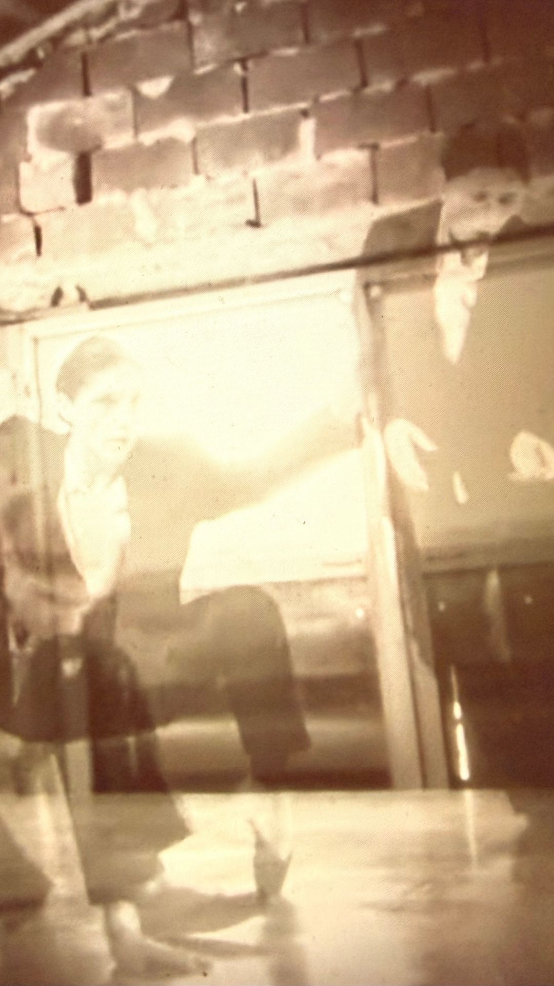 A sepia-toned image shows two ghostly figures superimposed against a brick wall, creating an eerie, mysterious atmosphere. A small, framed photograph of two people in dark attire hangs on the wall.