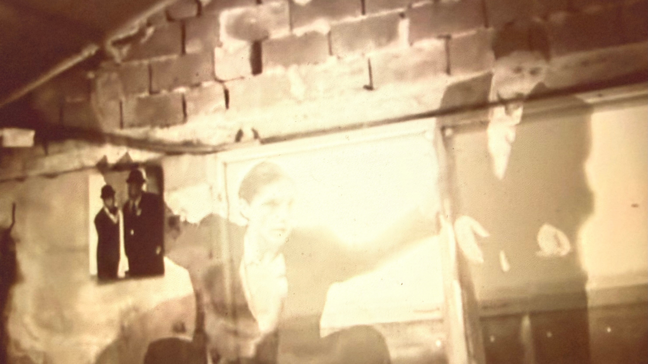 A sepia-toned image shows two ghostly figures superimposed against a brick wall, creating an eerie, mysterious atmosphere. A small, framed photograph of two people in dark attire hangs on the wall.