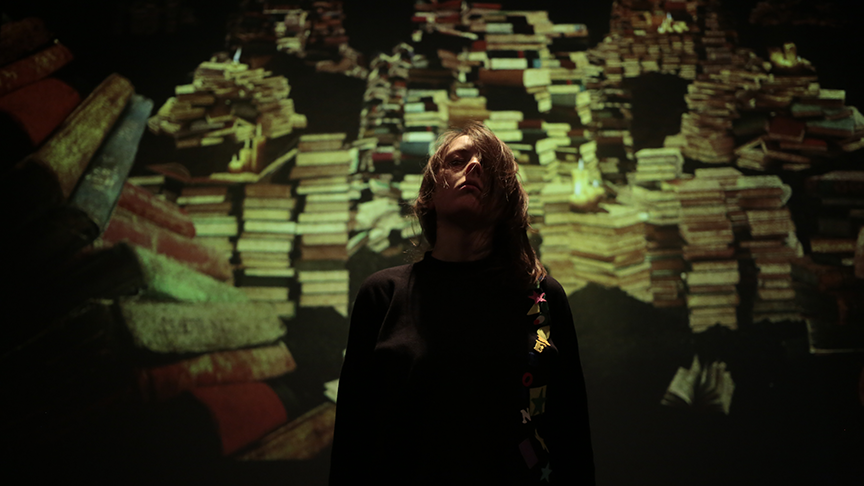 A person stands in shadow, illuminated by a warm, dim light, with an artistic backdrop of towering, colorful stacks of books creating a mysterious and introspective mood.