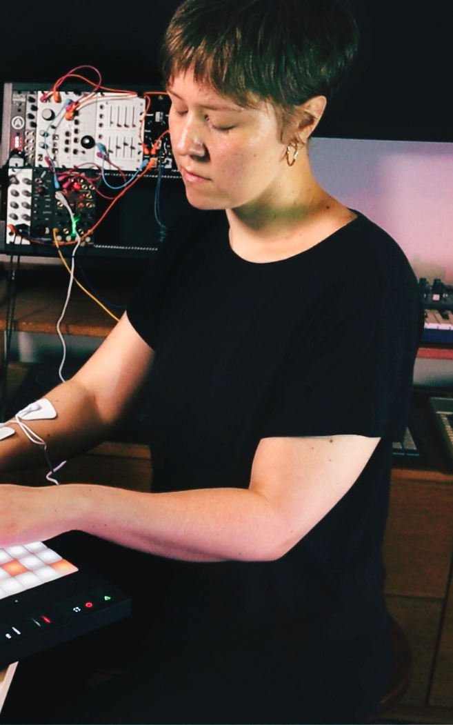 Liina, with a black top, short hair, and gold earrings is playing on a sample pad with electrodes connected to their arm.