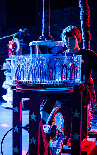 A dimly lit room with vibrant blue and red lighting. A person on the right pedals a bike that activates light-up figures. A smiling adult and child watch closely.