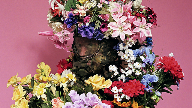 A person camouflaged in a vibrant array of flowers stands against a pink background. The mood is whimsical and colorful, blending nature and art.