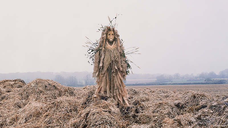 A figure stands in a misty field, wearing a costume of straw and branches. The scene is eerie and atmospheric, with muted tones and overcast sky.