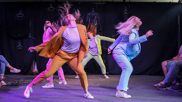 Four dancers in colorful suits perform energetically on stage with dynamic hair flips. Their lively movements and vibrant costumes create a sense of joy.