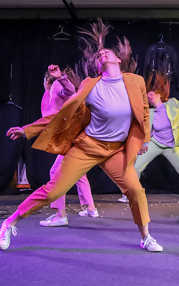 a dancer in a colourful suit performs energetically on stage with dynamic hair flips. Their lively movements and vibrant costumes create a sense of joy.